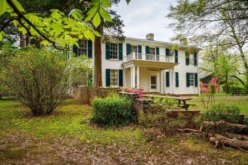 A historic white house with green shutters, surrounded by trees and blooming flowers in a serene garden setting.