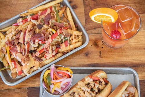 A tray of loaded fries topped with bacon and sauce, alongside a hot dog and a colorful cocktail with an orange slice.