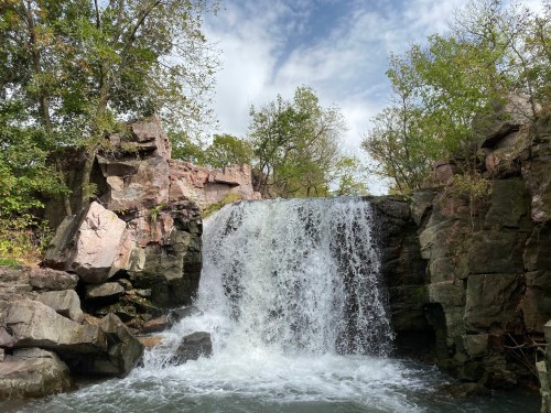A waterfall cascades over rocky cliffs, surrounded by lush greenery and a cloudy sky.