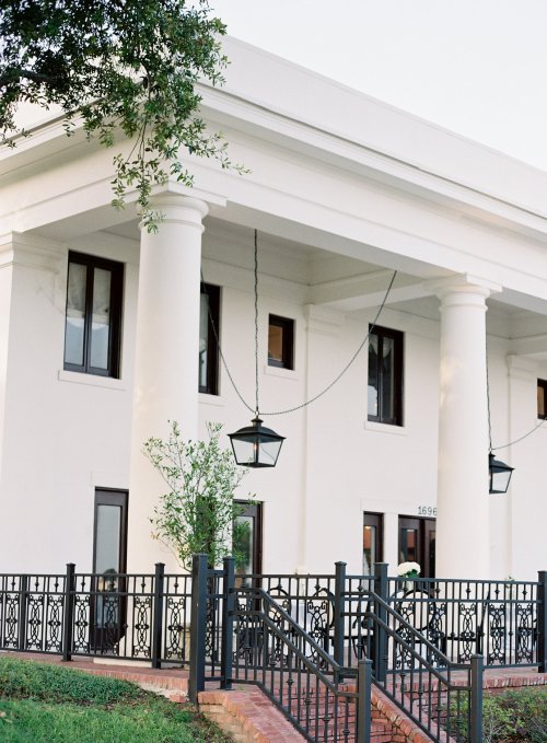 A white building with tall columns, large windows, and a decorative fence, surrounded by greenery.