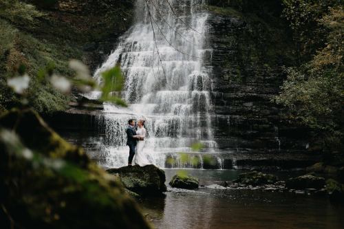 A couple embraces on a rock by a serene waterfall, surrounded by lush greenery.