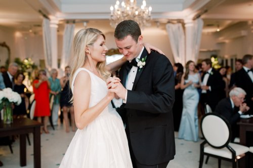 A couple dances together at a wedding reception, surrounded by guests in formal attire. Elegant decor in the background.