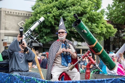 Two men in costumes, one in a wizard hat, hold telescopes on a festive parade float surrounded by greenery.