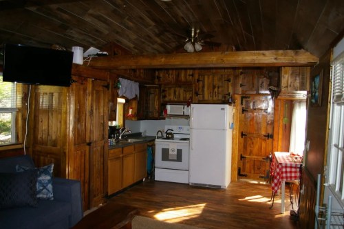 Cozy cabin interior featuring wooden walls, a kitchen with appliances, and a small dining area with a checkered tablecloth.