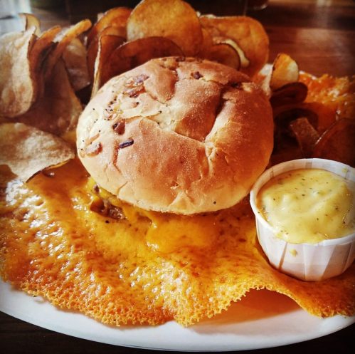A plate featuring a bun, crispy potato chips, and a small cup of dipping sauce on a cheesy, golden crust.