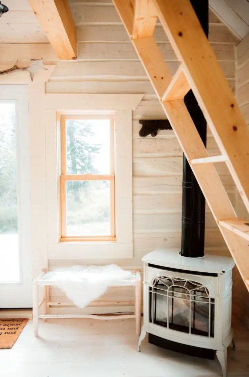 Cozy interior of a cabin featuring a wooden bench, a white stove, and a window with a view of nature.