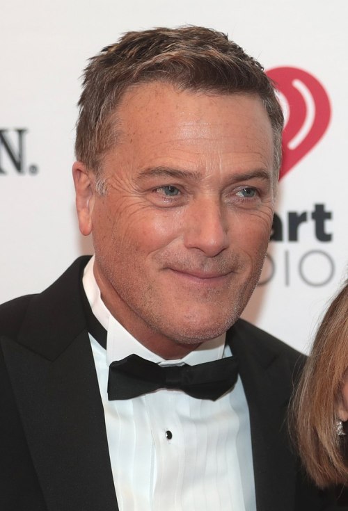 A man in a tuxedo smiles at an event, with a blurred background featuring logos and lights.