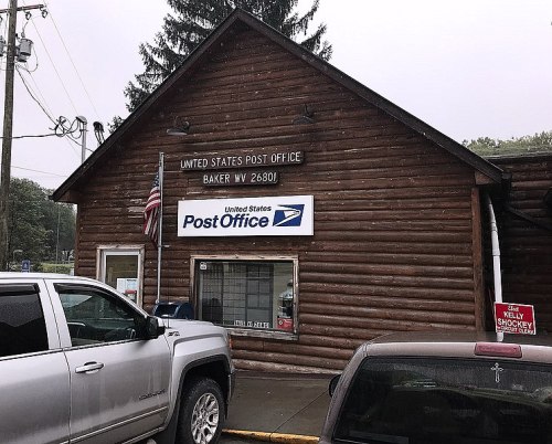 Exterior of a rustic log cabin post office with a U.S. flag, sign, and parked vehicles in front.