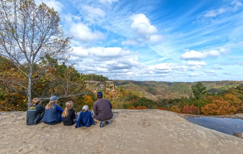 A family sits on a rocky ledge, overlooking a colorful autumn landscape with trees and a blue sky.