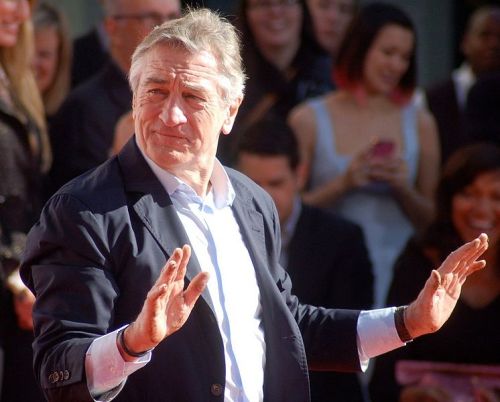 A man in a suit gestures with a smile, surrounded by a crowd at an outdoor event.