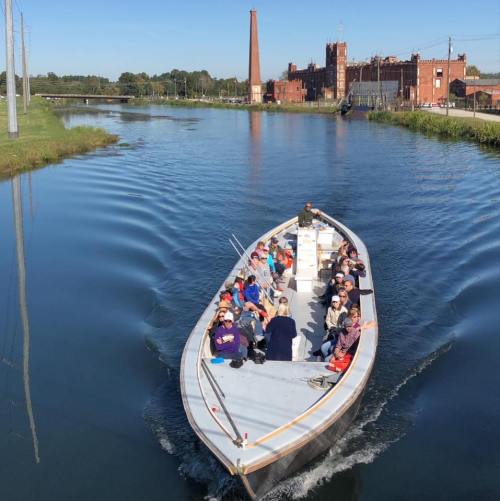 A group of people on a boat gliding through a calm canal, with a historic factory building in the background.