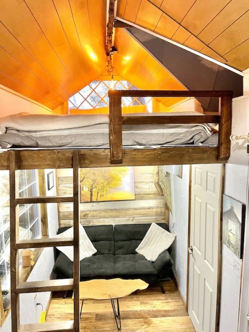 Cozy tiny home interior featuring a lofted bed, wooden ladder, and a green sofa beneath a warm, orange ceiling.