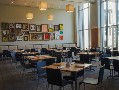 Modern restaurant interior with tables, chairs, and colorful artwork on the wall, featuring large windows for natural light.