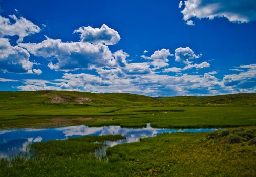 A serene landscape featuring green hills, a calm pond, and a bright blue sky with fluffy white clouds.