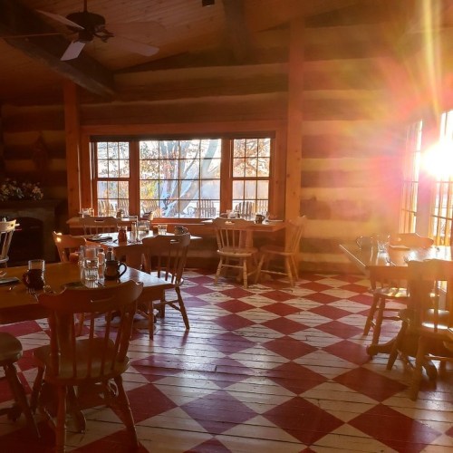 Cozy dining area with wooden tables and chairs, sunlight streaming through large windows, and a checkered floor.