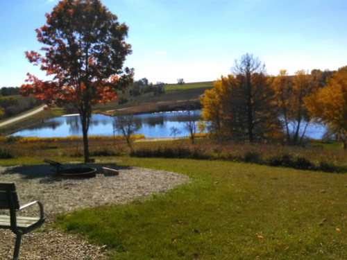 A serene landscape featuring a lake surrounded by autumn trees and a gravel area with a fire pit and bench.