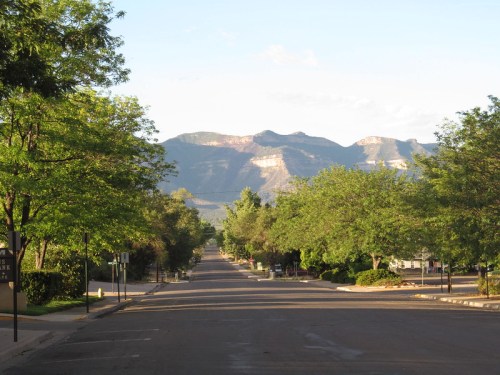 A quiet street lined with trees, leading to distant mountains under a clear blue sky.