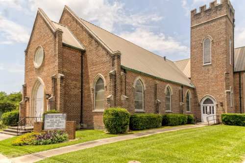 A brick church with arched windows and a tower, surrounded by green grass and colorful flower beds.