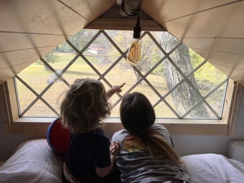 Two children sit on a bed, pointing out the window at a view of trees and a grassy yard. Natural light fills the room.