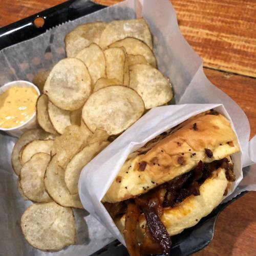 A sandwich with grilled onions in a bun, served with a side of crispy potato chips and dipping sauce.