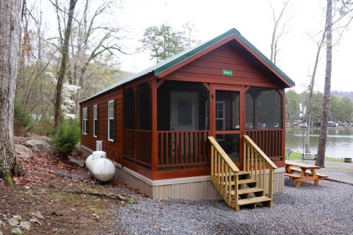 A cozy wooden cabin with a porch, surrounded by trees and near a lake, featuring a picnic table and a gas tank.