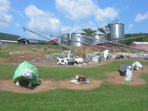 A farm scene featuring colorful rock sculptures in a grassy area, with silos and machinery in the background under a blue sky.