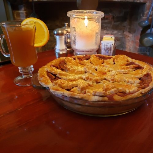 A slice of apple pie in a glass dish, a drink with a lemon slice, and a candle on a wooden table.