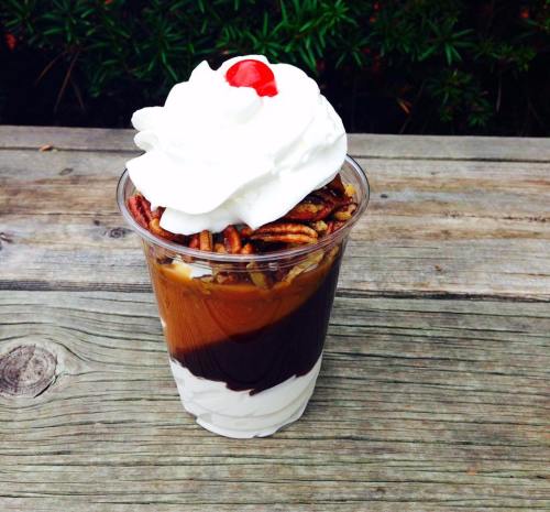 A dessert cup with layers of chocolate sauce, whipped cream, pecans, and a cherry on top, placed on a wooden surface.