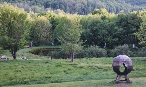 A serene landscape featuring a pond, lush greenery, and abstract sculptures among trees in a tranquil setting.
