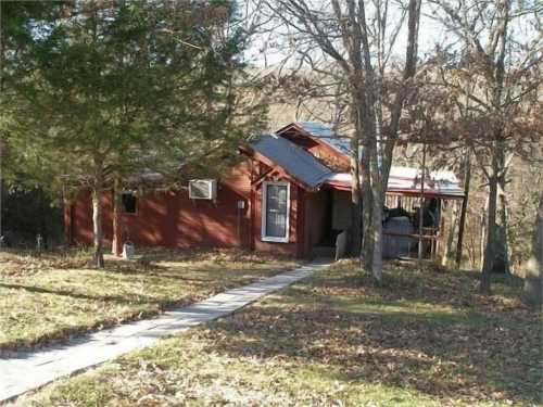 A small red cabin surrounded by trees, with a pathway leading to the entrance and a porch on the side.