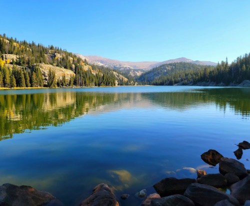 A serene lake surrounded by mountains and lush trees, reflecting the clear blue sky and calm water.