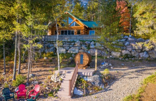 A log cabin on a hillside surrounded by trees, with a wooden deck and colorful chairs in the foreground.