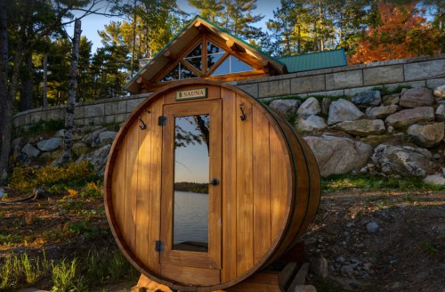 A wooden barrel sauna by a serene lake, surrounded by trees and a stone wall, with a cabin in the background.
