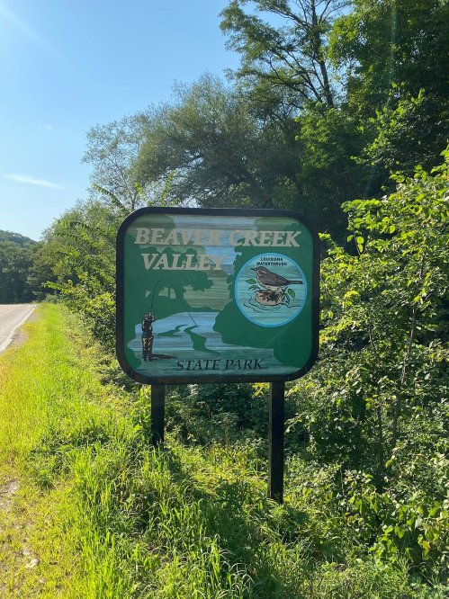 Sign for Beaver Creek Valley State Park, featuring a fishing scene and surrounded by greenery.