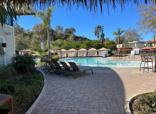 A sunny pool area with lounge chairs, cabanas, and palm trees, surrounded by greenery and a clear blue sky.