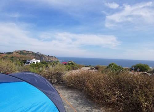 A view from a campsite with a blue tent, overlooking the ocean and distant hills under a partly cloudy sky.