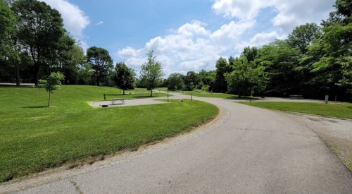 A winding path through a green park with trees and a picnic area under a partly cloudy sky.