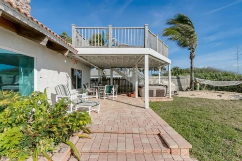 A sunny backyard with a patio, outdoor seating, a hot tub, and a deck overlooking a grassy area and palm trees.