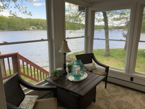 Cozy sunroom with lake view, featuring wicker furniture, a lantern, and decorative pillows, surrounded by greenery.