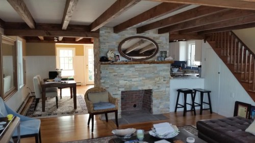 Cozy living room with a stone fireplace, wooden beams, and a view of a dining area and staircase.