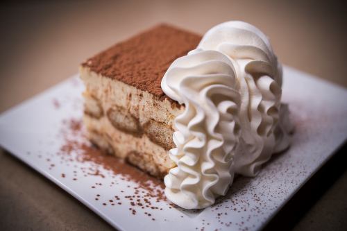 A slice of tiramisu topped with cocoa powder and a generous swirl of whipped cream on a white plate.