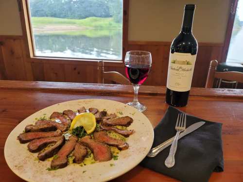 A plate of sliced steak with lemon, garnished with herbs, alongside a glass of red wine and a bottle on a wooden table.