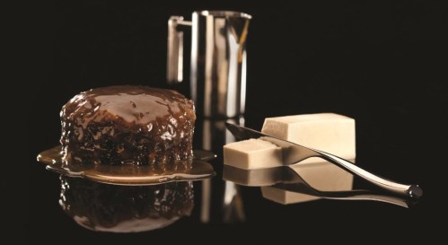 A round dessert with a glossy sauce, next to a knife and blocks of fudge, all set against a black background.