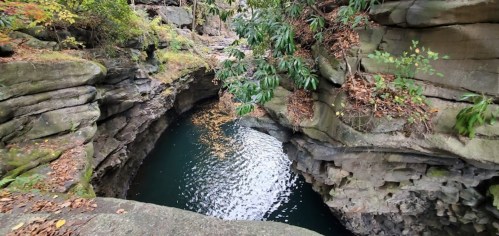 A serene, rocky gorge with clear water reflecting light, surrounded by lush greenery and autumn leaves.