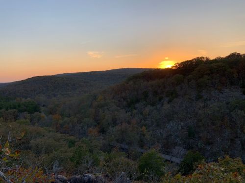 Sunset over a forested valley, with rolling hills and autumn foliage under a colorful sky.
