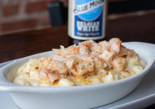 A creamy macaroni and cheese dish topped with shrimp, served in a white dish, with a beer bottle in the background.