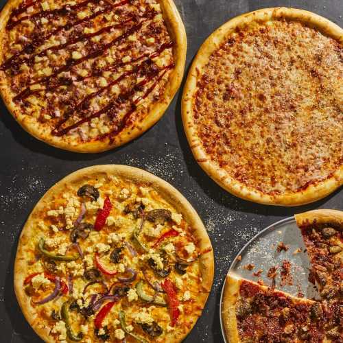 Four pizzas on a dark surface: BBQ chicken, cheese, veggie, and a meat pizza, with a slice removed from the meat pizza.