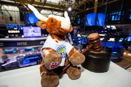 A plush bull wearing a shirt sits with a gavel in a stock exchange setting, with screens in the background.