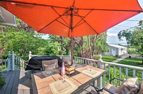 A patio table set with placemats under a large orange umbrella, surrounded by greenery and a grill in the background.