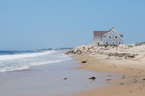 A beach scene featuring a gray house near the shore, with gentle waves and a clear blue sky in the background.